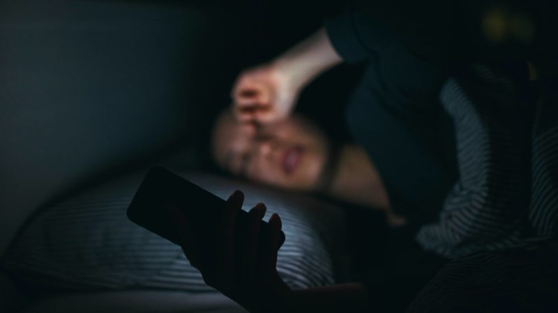 Tired woman rubbing eyes while using mobile phone on bed till late