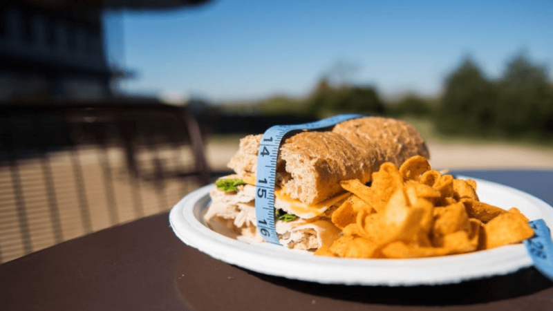 a sandwich and corn chips on a white plate. the sandwich has a tape measure around it
