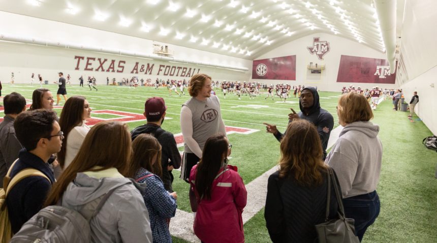 Aggie achieve students at a Texas A&M practice facility talking with football players