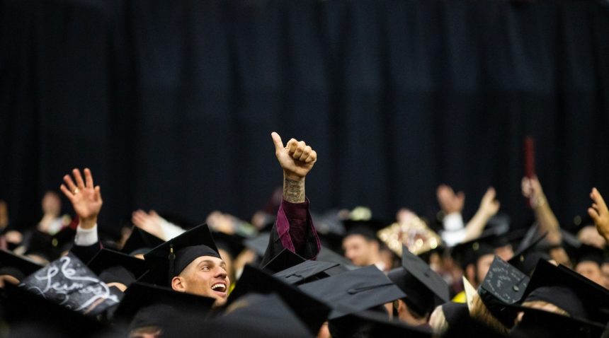 a student standing among graduations in caps and gowns flashes a gig 'em