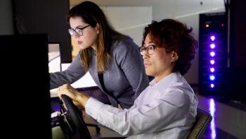 a woman observes a seated man using a virtual reality driving simulator