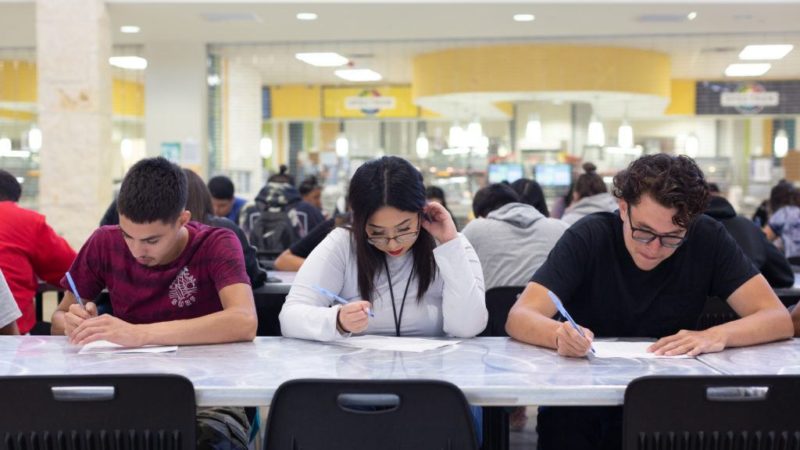 three students look down at papers while sitting at a cafeteria table