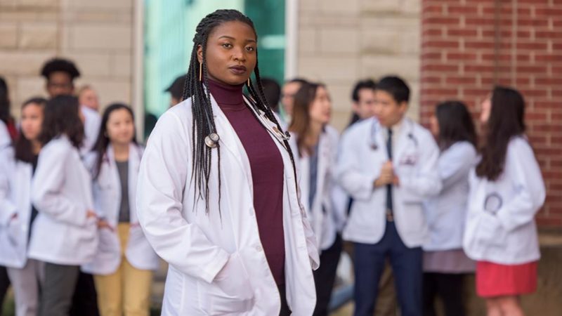 a black student in a white doctor's coat stands with her hands in her pockets