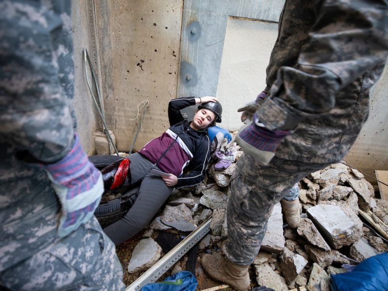 two men in army fatigues standing in front of a student lying in rubble at a mock earthquake site