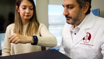 a woman holds up her wrist to look at a smartwatch, with a researcher looking on