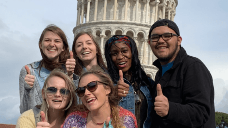 Six students pose with thumbs up in front of the Leaning Tower of Pisa