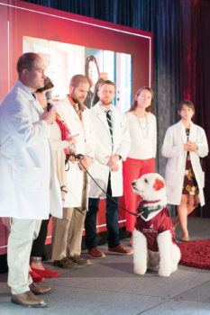 a dog seated on a stage in front of veterinarians in white coats