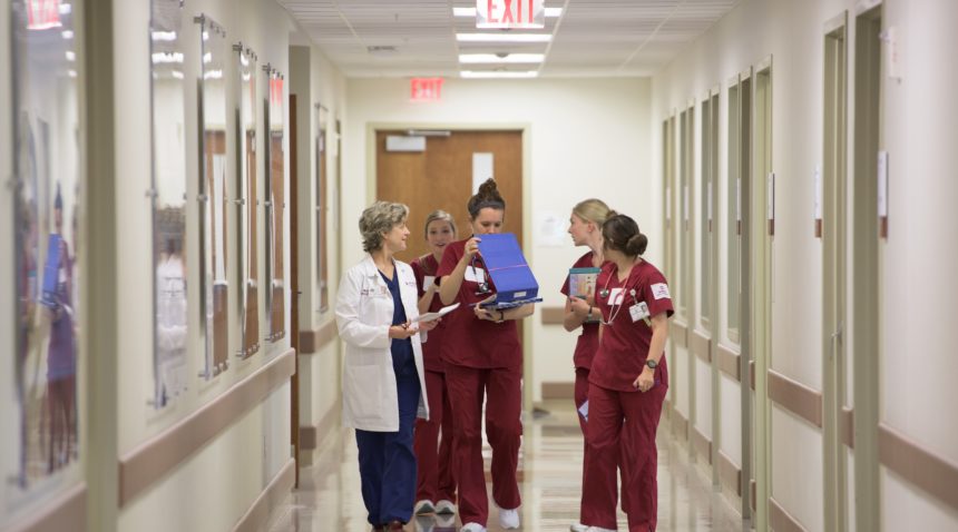 students in maroon scrubs and doctor in white lab coat walking down hall of hospital