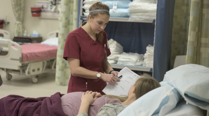 student in maroon scrubs in a hospital standing next to patient's bed
