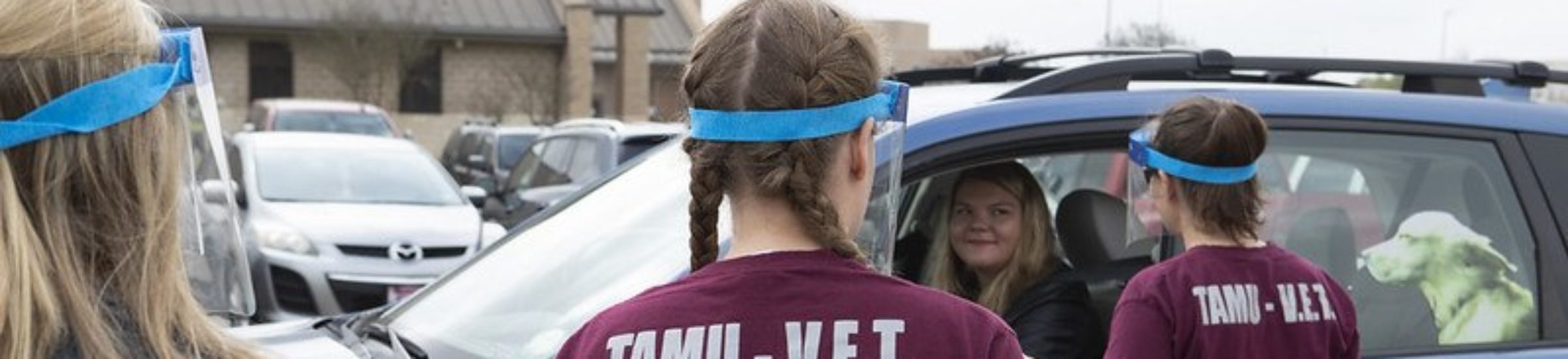 people wearing maroon vetmed shirts wearing protective masks talk to a woman in a car