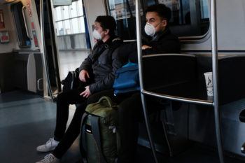 two men seated on an airport tram wearing protective face masks