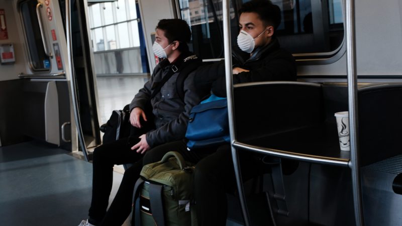 two men seated on an airport tram wearing protective face masks