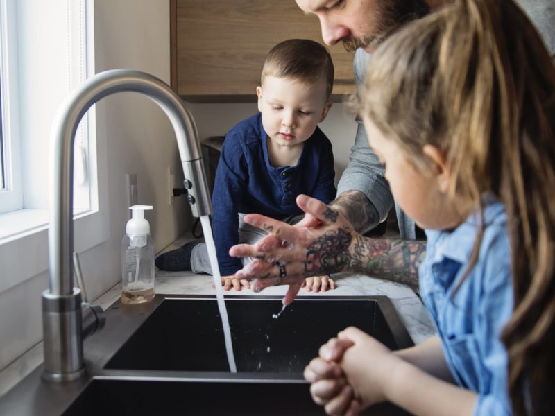 father washing hands at sink while toddler watchers on counter and older child leans on sink