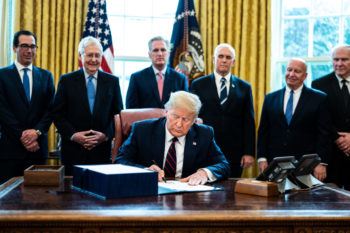 president donald trump seated in the oval office signing bill while cabinet members look on