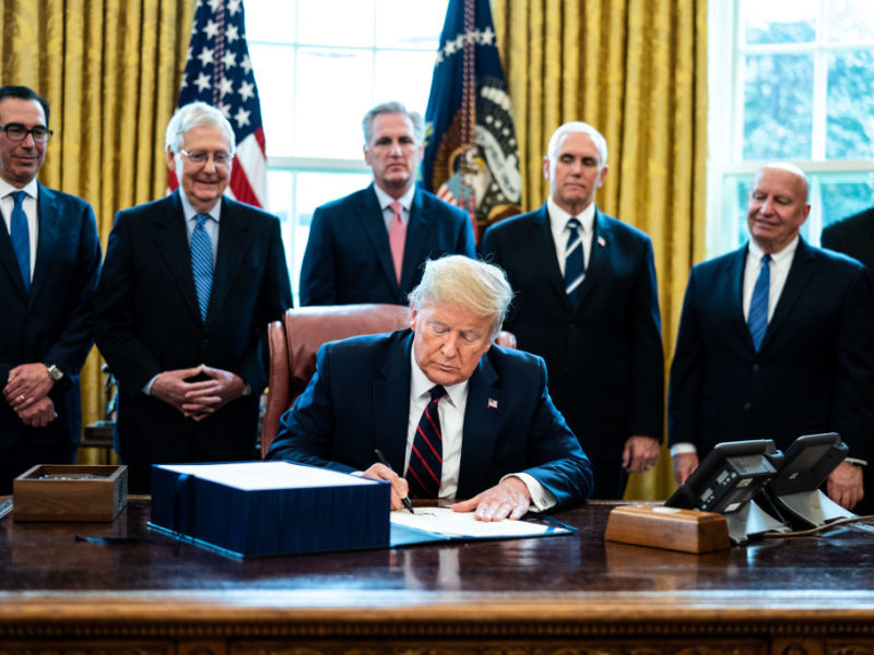president donald trump seated in the oval office signing bill while cabinet members look on