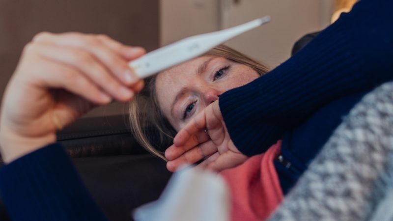 an ill woman on a couch looking at a thermometer