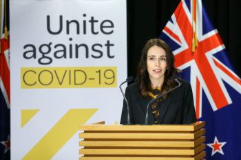 jacinda ardern speaking at a podium in front of a "unite against covid-19" sign and a new zealand flag