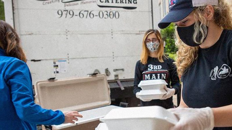 women wearing masks and gloves handle boxed meals with graphic overlay reading #support aggieland