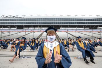 student wearing graduation cap and gown with face mask sitting in foreground in front of other students spaced six feet apart in the empty texas motor speedway