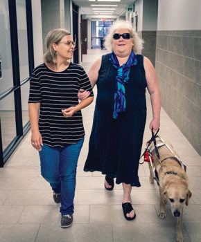 a woman standing to the left holds the arm of a woman who is blind while walking down the hallway aloong with a service dog