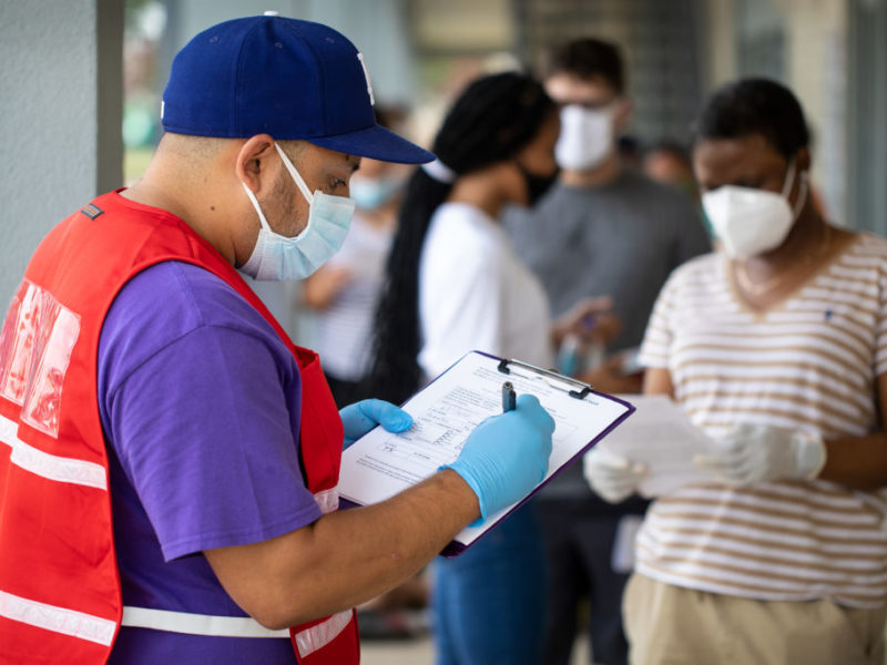 man wearing masks and gloves writes on a clipboard in front of a line of people
