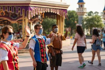 disney cast members stand in theme park wearing face masks welcoming guests