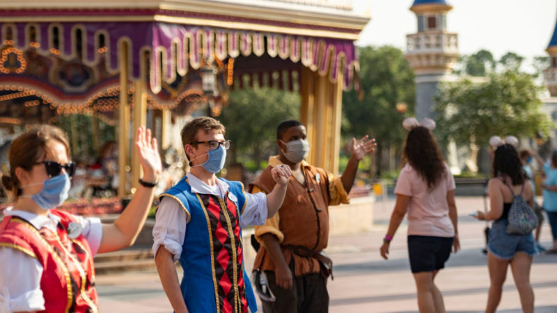 disney cast members stand in theme park wearing face masks welcoming guests