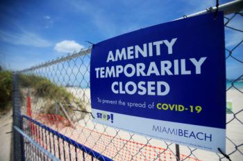 close up of a sign on a fence saying "menity temporarily closed" with beach in backround