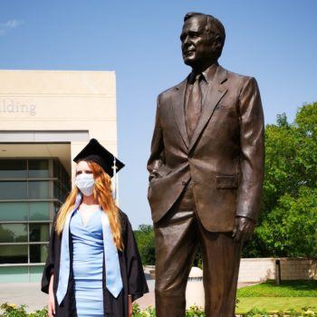 student in cap and gown wearing face covering standing next to george h.w. bush statue