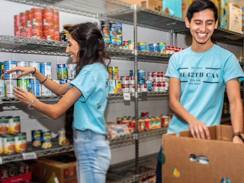two students wearing blue t shirts standing in food pantry filled with canned goods