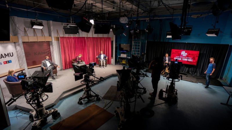 view of cameras and lights pointing toward stage, with three seated peopl being interviewed