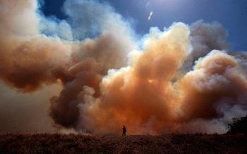 a firefighter stands in the distance of a field against a large plume of smoke in the sky