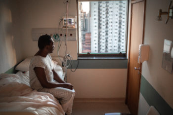 black man in hospital gown sitting on bed in hospital and looking out window