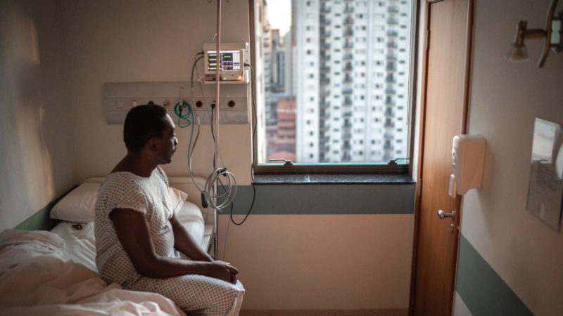 black man in hospital gown sitting on bed in hospital and looking out window