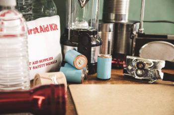 A pile of emergency preparation, natural disaster supplies including: flashlight, first aid kit, lantern, water bottles, canned goods, can opener, radio, backpack, batteries.