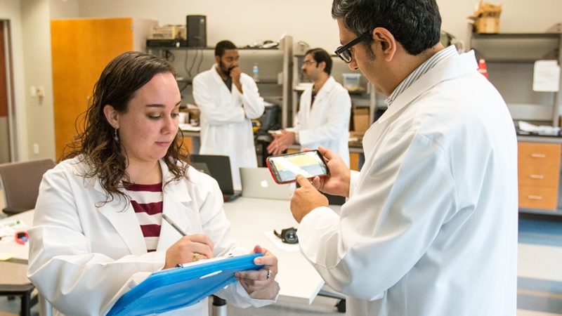 two researchers in white coats work in a lab with two other researchers in the background