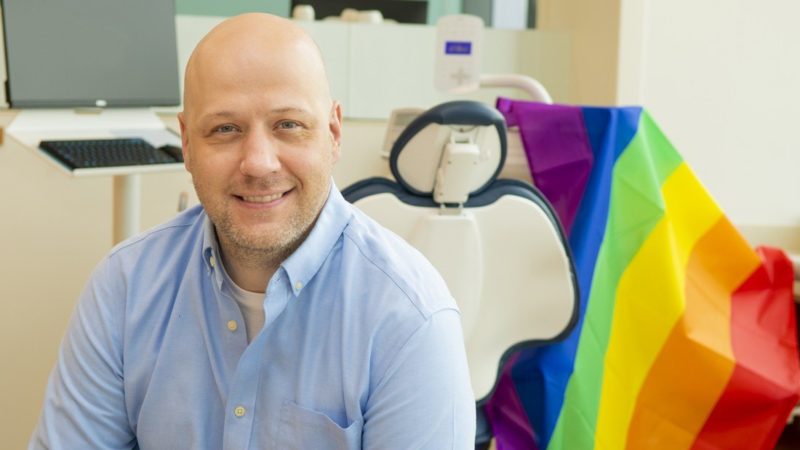 Matthew Kesterke sitting in dental chair draped with rainbow Pride flag