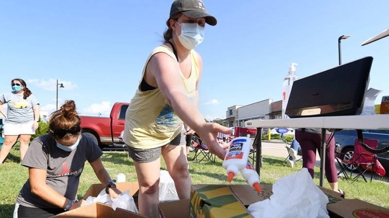support aggieland text on a photo of a woman wearing a mask tossing bottles of glue into a box of school supplies