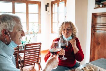 two elderly people wearing face masks cheers with wine glasses