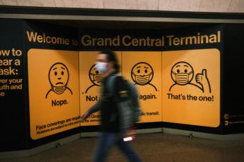 blurred image of man wearing face mask walking by display in grand central terminal displaying how to properly wear a face mask