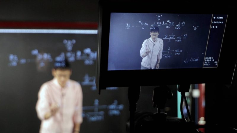 a camera's viewfinder focused on a mathemetician in front of a blackboard covered in equations