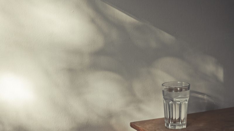 Glass of water on wooden table next to wall with shadows coming from window.