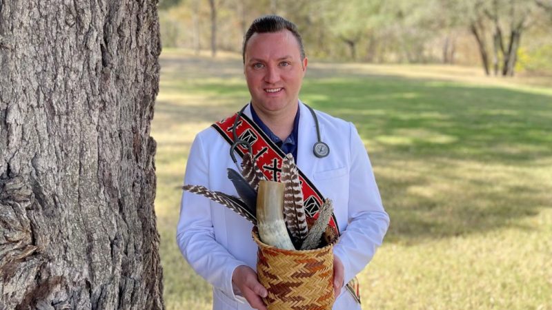 bruce cody standing outside in his white nurse's coat wearing his traditional Native American regalia