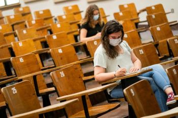 two students wearing face coverings sit at desks in an empty lecture hall