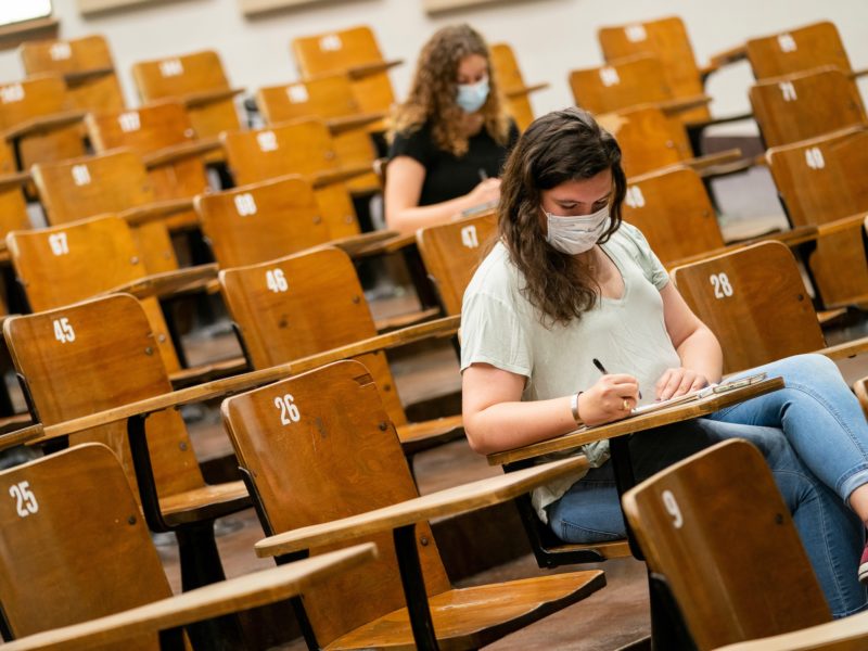 two students wearing face coverings sit at desks in an empty lecture hall