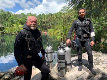 Iliffe and Calderon stand side by side in wet suits with oxygen tanks near the water in a tropical setting
