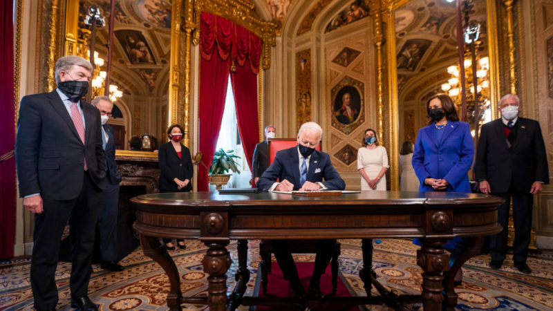 joe biden seated at desk siging documents surrounded by officials and kamala harris
