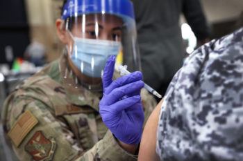 A woman in national guard fatigues wearing a face shield administers a vaccine