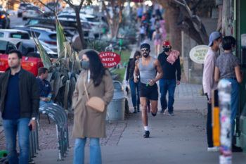 pedestrians walk down a commercial street, some wearing face masks