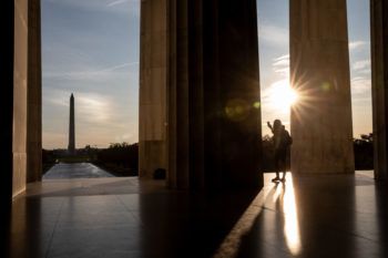 a woman is backlit by the sun as she takes a photo of the washington monument from the lincoln memorial